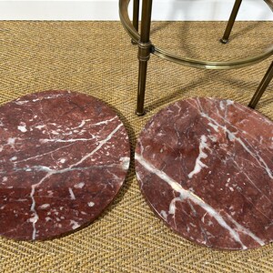 May include: Two round, red marble table tops with white veining are displayed. The marble has a polished finish. A brass-colored metal bar stool is partially visible in the background.