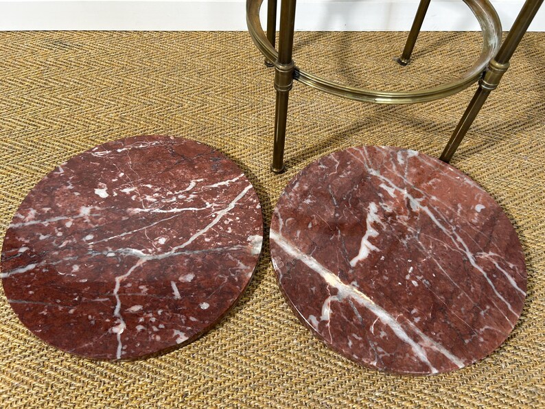 May include: Two round, red marble table tops with white veining are displayed. The marble has a polished finish. A brass-colored metal bar stool is partially visible in the background.