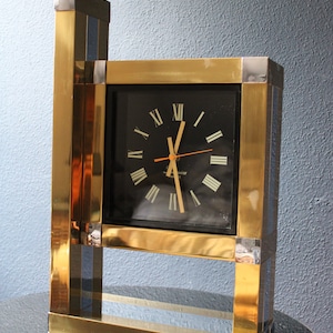 May include: A gold-toned clock with Roman numeral markings on a black face. The clock is framed in a gold metal frame with silver accents. The clock is standing on a black surface.