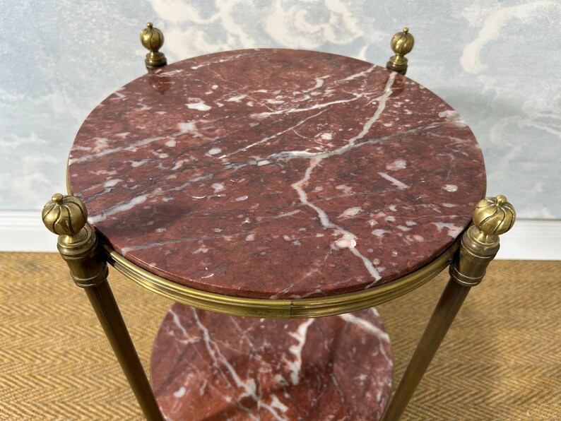 May include: A small, round side table with a red marble top and a lower shelf. The table has a brass frame and decorative finials. The marble has white veining. The table is approximately 24 inches in diameter.