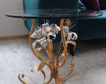 May include: A gold metal side table with a round glass top. The table features a decorative design of stylized flowers and leaves.