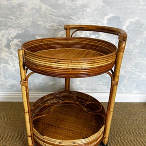 May include: A two-tiered, vintage-style bamboo bar cart. The cart features two circular trays with woven rattan bases and a handle. The frame is made of bamboo, with brass-colored accents on the feet. The cart is designed for serving drinks and entertaining.