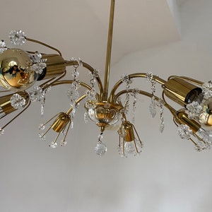 Op de afbeelding: Een gouden kroonluchter met meerdere armen en kristallen accenten. Elke arm heeft een heldere glazen lamp en een decoratief kristalbloemmotief.