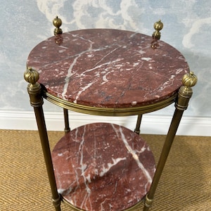 May include: A small, two-tiered side table with a round, red marble top and a lower shelf. The marble has white veining. The table has brass accents and legs, and decorative finials. The table is approximately 24 inches tall.