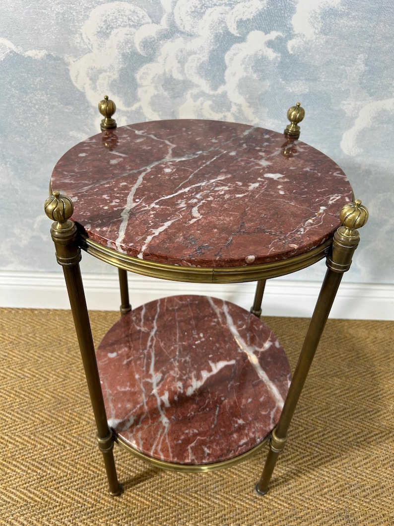 May include: A small, two-tiered side table with a round, red marble top and a lower shelf. The marble has white veining. The table has brass accents and legs, and decorative finials. The table is approximately 24 inches tall.