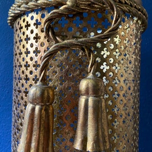 May include: Gold metal lampshade with a decorative rope design and two tassels. The lampshade has a perforated pattern.