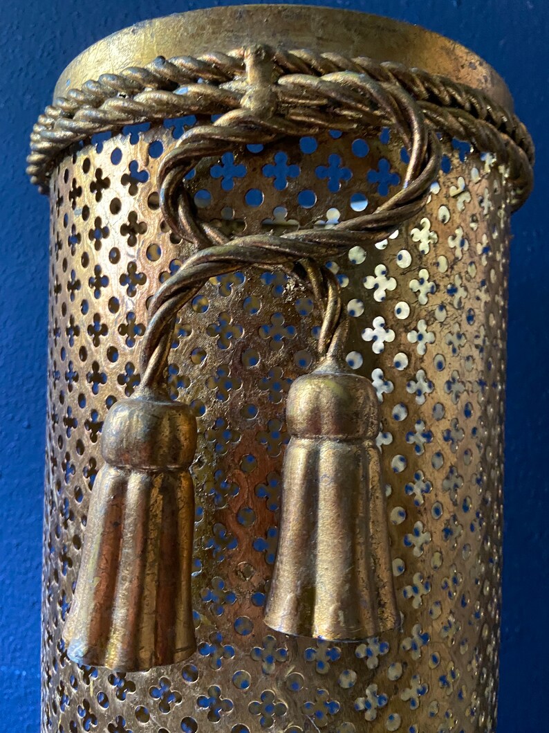 May include: Gold metal lampshade with a decorative rope design and two tassels. The lampshade has a perforated pattern.