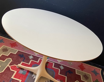 May include: White oval table with a white pedestal base and four legs. The table is on a red, pink, and blue patterned rug.