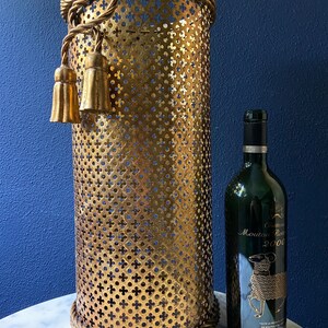 May include: Gold metal umbrella stand with a rope design around the top and bottom. The stand has a decorative tassel on the side. A bottle of Chateau Mouton Rothschild 2000 wine is next to the stand.