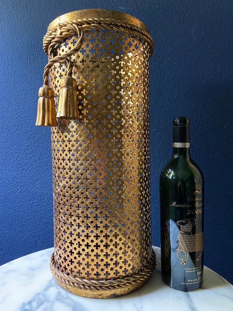May include: Gold metal umbrella stand with a rope design around the top and bottom. The stand has a decorative tassel on the side. A bottle of Chateau Mouton Rothschild 2000 wine is next to the stand.