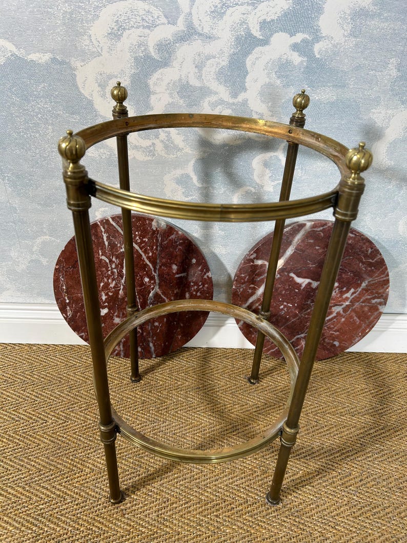 May include: A vintage brass side table with a circular top and base, supported by four slender legs. The table features two round, red marble inserts. The design includes decorative finials at the top.
