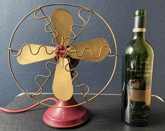 May include: A vintage red and gold Siemens fan with a metal cage and three blades. The fan is sitting on a table next to a bottle of Chateau Mouton Rothschild 2000 wine.