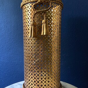 May include: Gold metal umbrella stand with a decorative rope design and tassels. The stand has a perforated design.