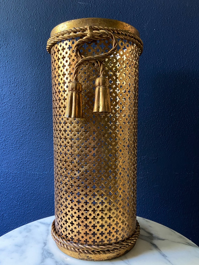 May include: Gold metal umbrella stand with a decorative rope design and tassels. The stand has a perforated design.