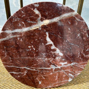 May include: A round table top made of red marble with white veining. The marble has a polished finish and is supported by brass legs. The table is a decorative accent piece.