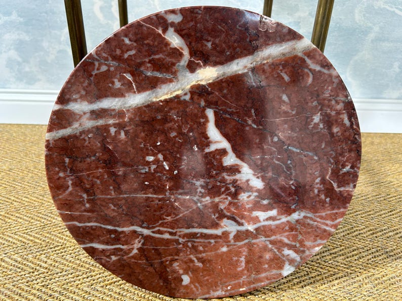 May include: A round table top made of red marble with white veining. The marble has a polished finish and is supported by brass legs. The table is a decorative accent piece.