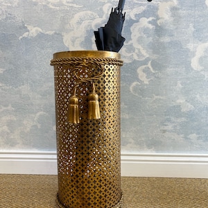 May include: A gold metal umbrella stand with a decorative rope design around the top and bottom. The stand has a perforated design and two tassel accents. A black umbrella is inside the stand.