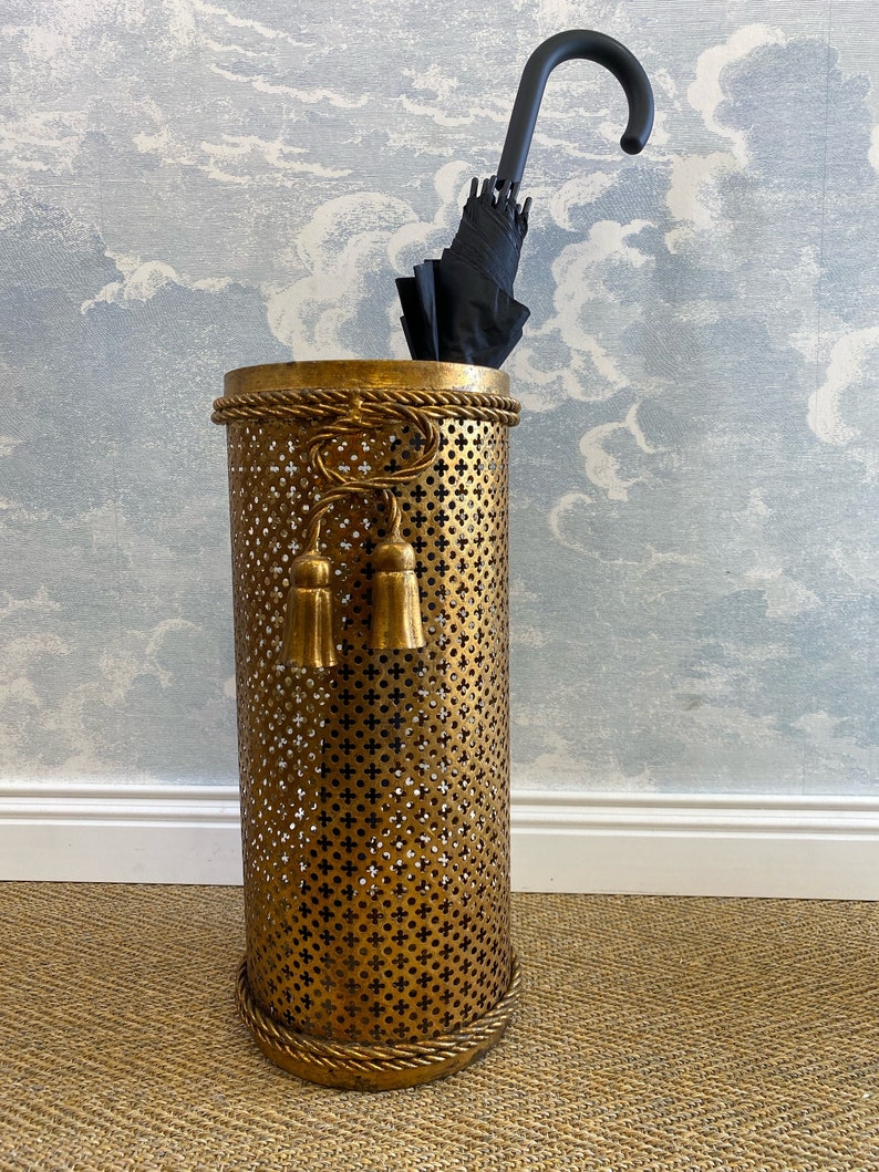 May include: A gold metal umbrella stand with a decorative rope design around the top and bottom. The stand has a perforated design and two tassel accents. A black umbrella is inside the stand.