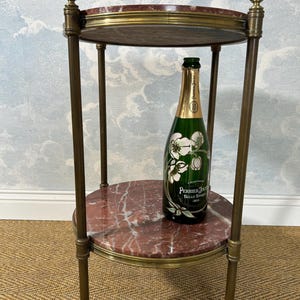 May include: A two-tiered side table with a red marble top and brass accents. A green champagne bottle with white floral graphics sits on the lower tier. The table has a classic, elegant design.