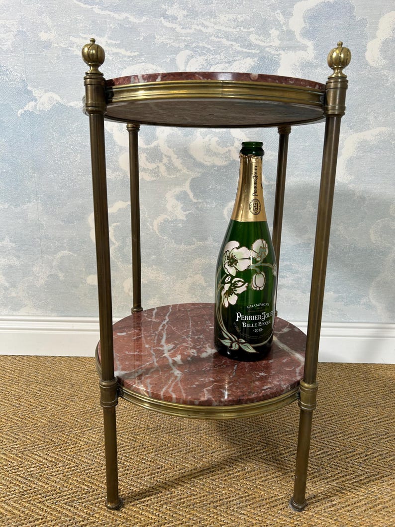 May include: A two-tiered side table with a red marble top and brass accents. A green champagne bottle with white floral graphics sits on the lower tier. The table has a classic, elegant design.