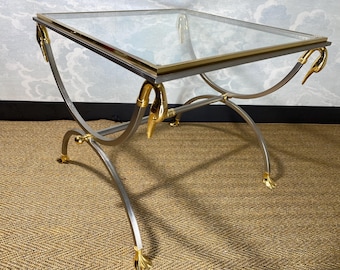 May include: A glass-top side table with a silver metal frame and gold swan accents. The table has a curved design and sits on a brown woven rug.