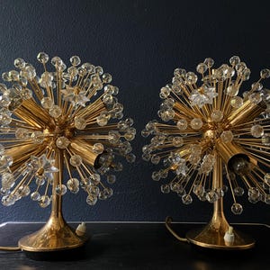 Op de afbeelding: Een paar gouden tafellampen met een uniek ontwerp met een bol van kristal-achtige accenten. De lampen hebben een gouden voet en een gouden snoer.