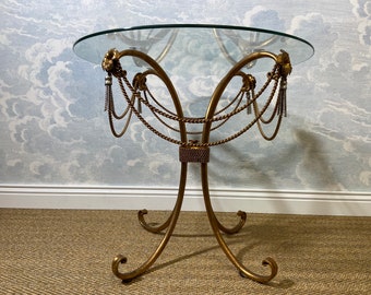 May include: A gold metal table with a glass top. The table has a decorative design of rope and tassels.