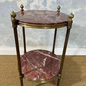 May include: A small, two-tiered side table with a red marble top and a lower shelf. The table has a brass frame and decorative finials. The marble has white veining. The table is approximately 24 inches tall.