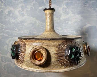 May include: A ceramic pendant light fixture with a brown and beige finish. The fixture has a round, flat base with a central dome and several decorative accents. The accents include small, round, colored glass inserts and raised, textured designs.