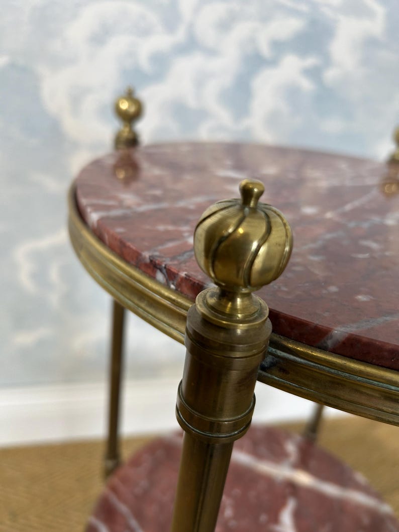 May include: A small, antique side table with a red marble top and brass accents. The table features a rounded oval top, supported by brass legs and decorative finials. The marble top has a reddish-brown color with white veining.
