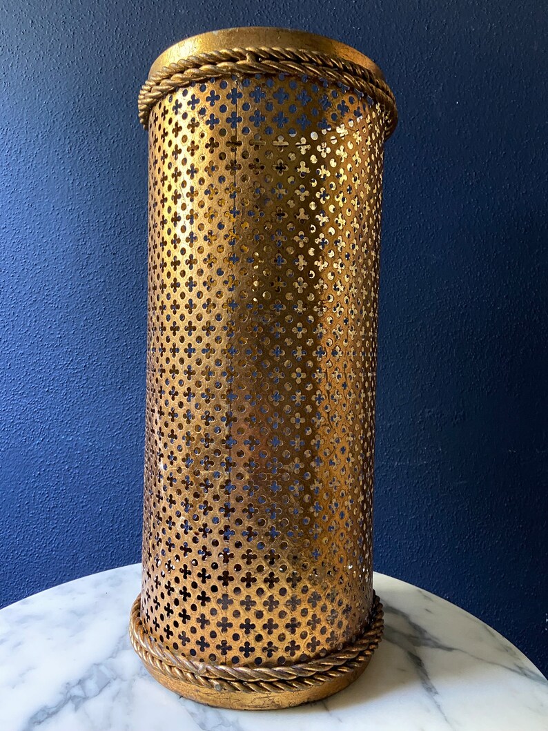 May include: Gold metal umbrella stand with a decorative rope trim and a perforated design.