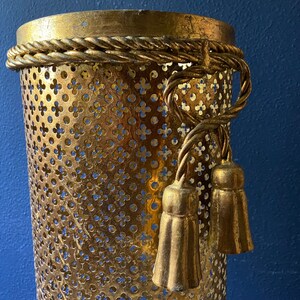 May include: Gold metal umbrella stand with a decorative rope trim and tassels. The stand has a perforated design.