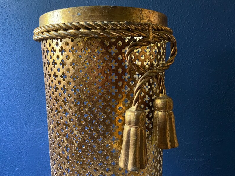 May include: Gold metal umbrella stand with a decorative rope trim and tassels. The stand has a perforated design.