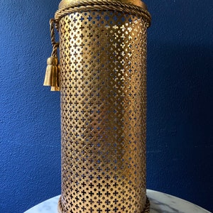 May include: Gold metal umbrella stand with a decorative rope trim and a tassel. The stand has a perforated design.