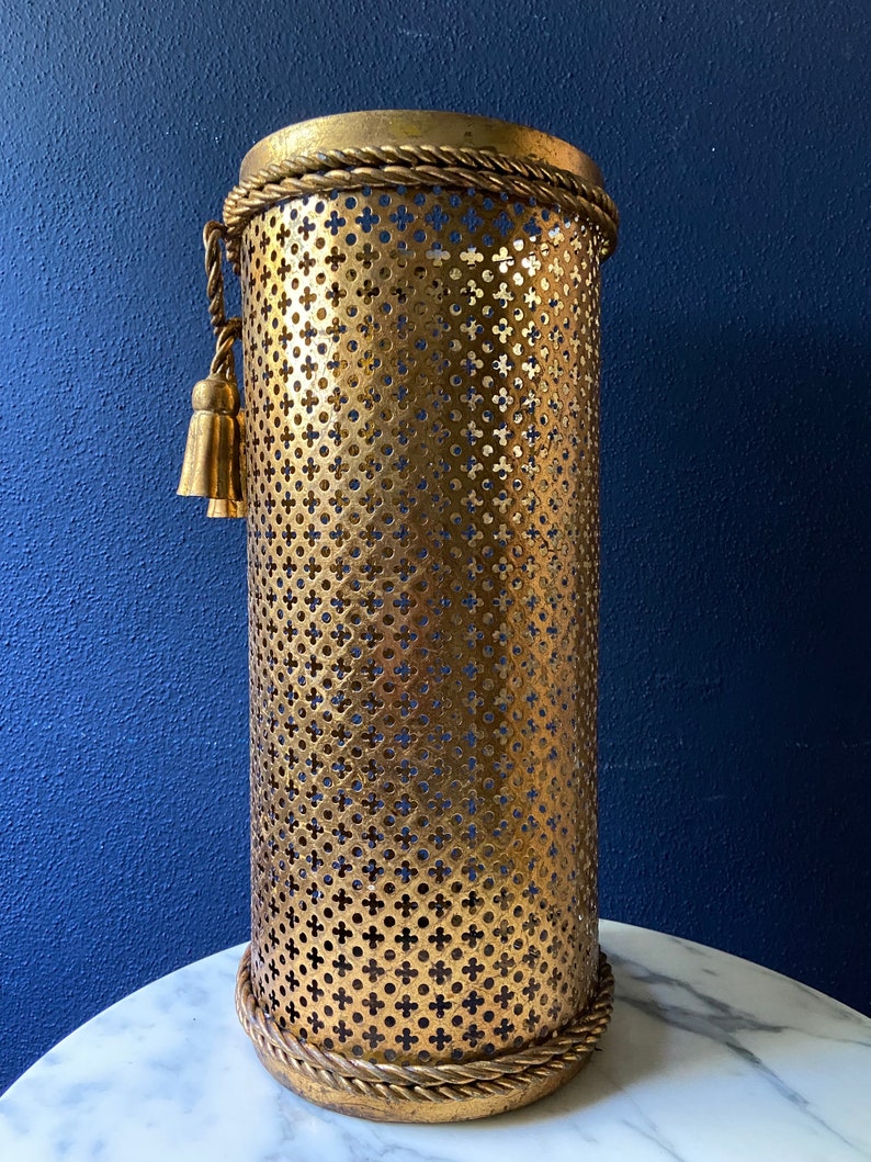 May include: Gold metal umbrella stand with a decorative rope trim and a tassel. The stand has a perforated design.