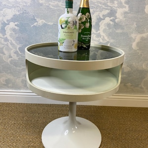 May include: A white, mid-century style side table with a round top and a pedestal base. The table has a dark glass top and a lower shelf. Two bottles of alcohol are on the table. One is a green bottle of champagne, the other a white gin bottle.