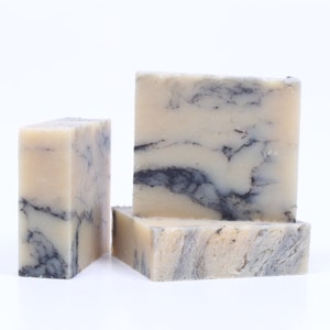 May include: Three rectangular bars of soap with a marbled design. The soap has a light beige base color with dark gray and black veining throughout. The bars are stacked and arranged on a white surface.