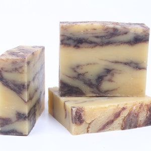 May include: Four rectangular bars of handmade soap. The soap has a light beige base color with dark brown marbling throughout. The bars are stacked and arranged on a white background.