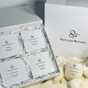 May include: A white gift box filled with four small white boxes containing soy candles. Each box has a black butterfly logo and the text "Papillon Natural" with the candle scent name. The box is sitting on a white fluffy surface. A fifth candle in a clear glass jar with a black butterfly logo and the text "Papillon Natural" with the candle scent name is sitting on the fluffy surface.