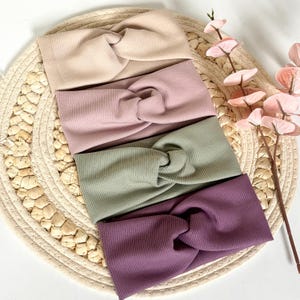 Könnte beinhalten: Ein Set aus vier breiten Stirnbändern aus geripptem Stoff, die jeweils ein zentrales Knotendetail aufweisen. Die Farben umfassen Beige, Altrosa, Salbeigrün und Dunkelpflaume. Die Stirnbänder sind in einem vertikalen Stapel angeordnet.
