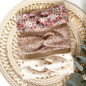 Könnte beinhalten: Drei gerippte Stoffstirnbänder mit gedrehtem Vorderteil. Das obere Stirnband zeigt ein dichtes Blumenmuster in Rot-, Rosa- und Grüntönen. Das mittlere Stirnband hat einen pfirsichfarbenen Blumendruck. Das untere Stirnband ist cremefarben mit kleinen Blumen.