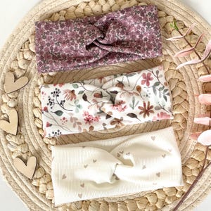 Könnte beinhalten: Drei gerippte Stirnbänder mit gedrehtem Vorderteil. Das obere Stirnband hat ein dunkelmalvenfarbenes Blumenmuster, das mittlere Stirnband eine weiße Basis mit buntem Blumendruck und das untere Stirnband ist cremefarben mit kleinen braunen Herzen.
