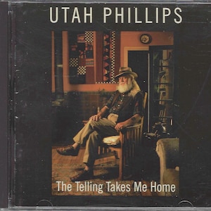 May include: A black and white CD case with a photo of a man with a long white beard wearing a hat and sitting in a chair. The text on the case reads "Utah Phillips" and "The Telling Takes Me Home".