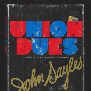 May include: A vintage book cover for "Union Dues" by John Sayles. The title is in bold red and blue letters against a distressed black background. The author's name is in gold script.