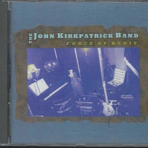 May include: A CD case with a blue background and a faded brown border. The text "The John Kirkpatrick Band Force of Habit" is printed in white on the front of the case. The image shows a blurry photo of a band in a recording studio.
