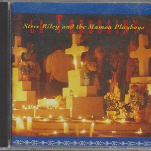 May include: CD case featuring the album art for "La Toussaint" by Steve Riley and the Mamou Playboys. The artwork depicts a nighttime scene with lit candles, crosses, and figures, creating a warm, atmospheric glow. The album title and artist name are displayed in gold lettering.