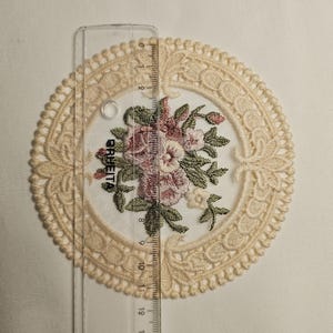 May include: A cream-coloured lace doily with an embroidered floral design in pink, green, and white. The doily is approximately 13 cm in diameter. A clear ruler is placed over the doily.