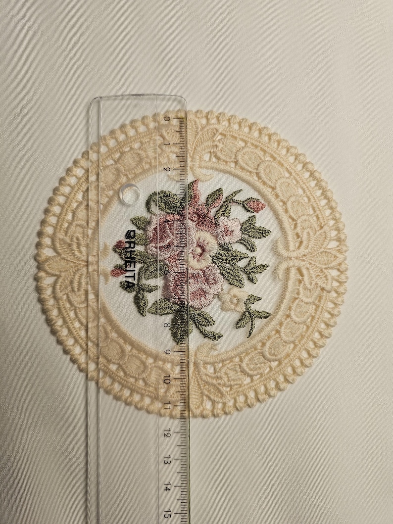 May include: A cream-coloured lace doily with an embroidered floral design in pink, green, and white. The doily is approximately 13 cm in diameter. A clear ruler is placed over the doily.