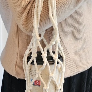 以下が含まれることがあります： 白のマクラメコーヒーカップホルダー、水色のハンドル付き。使い捨てコーヒーカップを持ち運ぶように設計されています。カップはベージュで、黒い蓋が付いています。ホルダーは肩からかけています。