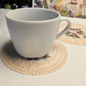 May include: A white ceramic teacup rests on a beige lace doily with a floral pattern. The doily is on a white surface. The background features a colourful illustration and blurred lights.